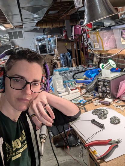 A selfie of a white woman with purple headphones on, in an electronics workshop, with some circular electronic boards and a battery holder on the table in front of her. She is casually holding a soldering iron and resting her head on her hand.
