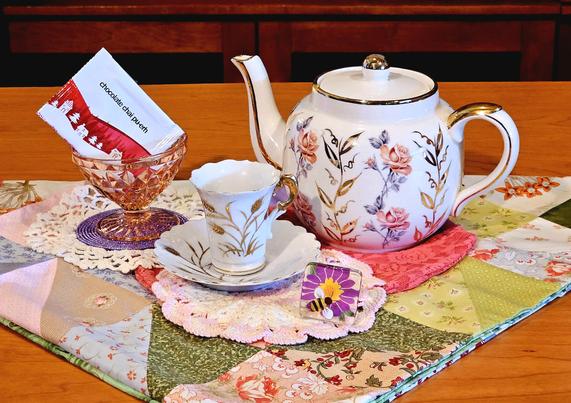 A tea tableau in summery colors of peach, yellow, green, orange, and purple. There is pink Depression glass, vintage china with gleaming gold leaf trim, a cheery honeybee painted on a glass flower, and vintage crocheted doilies, all resting upon a zestfully- colored quilted square. The tea is Chocolate Chai Puerh from