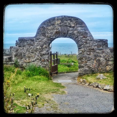 An old stone arch forming a gateway on a path to a church. The wooden gate is open and a path amongst the grass goes through the arch and to the right. The sea is visible in the background through the arch.