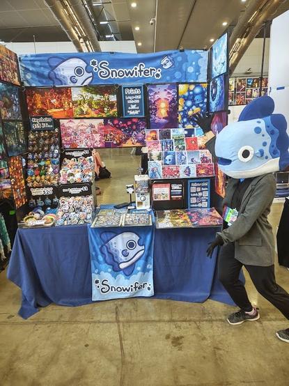 An artist alley table with colorful art. There is a person with a mola for a head showing off the booth.