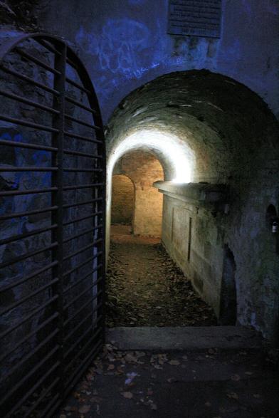 An archway set in a wall, at night. To the left is the metal gate that is usually locked. A series of horizontal bars set in a arched frame.
Inside, the tunnel is arched. The visible sideway visible is constructed from sandstone blocks. There's a ledge about 2/3 of the way up, then the wall curves over to become the roof. A light shines up the wall and spreads out across the roof. Beyond, is another wall with an arched doorway. A second passage runs off to the right.