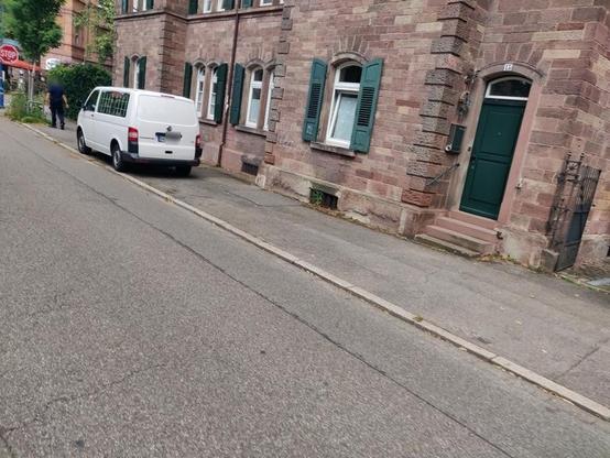 Einer weißer Bulli vom Land parkt auf dem Gehweg einer Nebenstraße. Ein Sansteinhaus säumt den Gehweg.