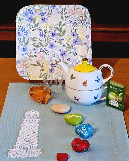 A tea tableau on a pale aqua mat showing a path made of hearts in various colors & materials, next to a lace lighthouse, leading toward a flowery meadow. There is a butterfly teapot & cup, and the tea is Green tea from