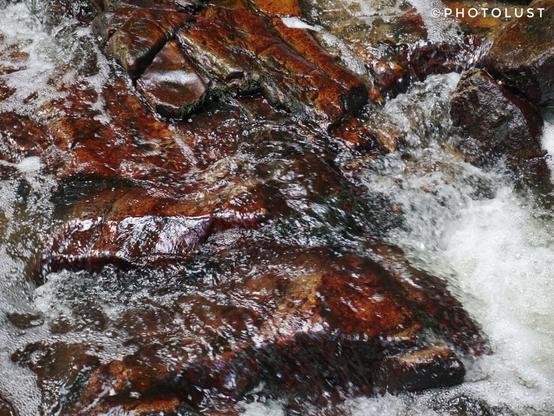 Wildwasser fließt über glatt geschliffene Steine mit rot-brauner Oberfläche, die in einem Bachbett liegen.