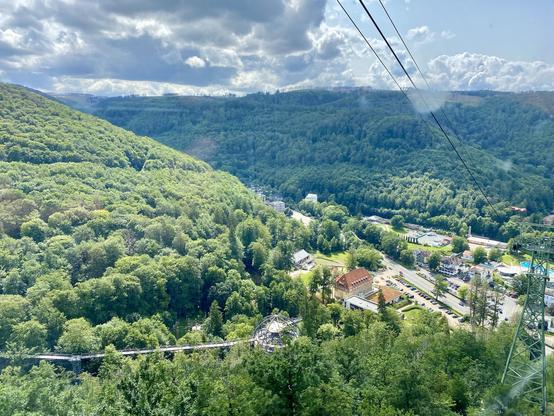 Ein malerischer Blick auf ein üppiges grünes Tal mit dichten Wäldern, einer kleinen Stadt und einer kurvenreichen Straße. Stromkabel verlaufen über das Bild und deuten auf eine nahe gelegene Seilbahn oder Gondel hin. Der Himmel ist teilweise bewölkt, was der Landschaft Tiefe verleiht.