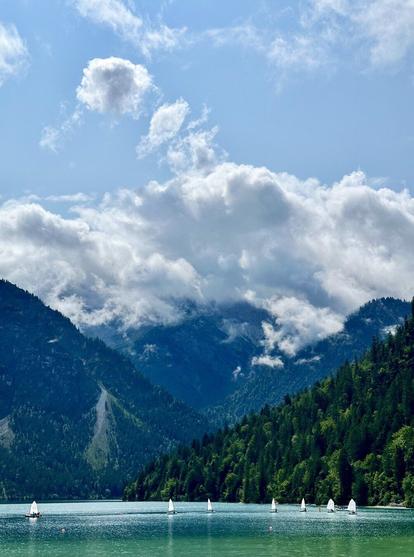 A serene lake surrounded by forested mountains with sailboats on the water and clouds in the sky.