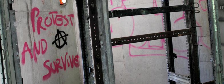 Im Vordergrund sieht man einen Ausschnitt eines Metallregals. Vom Regal selbst sieht man nur die Streben. Die Böden fehlen. Zwischen den Metallstäben sieht man auf einer Betonwand im Hintergrund Graffiti. Der Schriftzug "Protest and survive" ist mit einem Anarchie-Zeichen deutlich zu erkennen.
