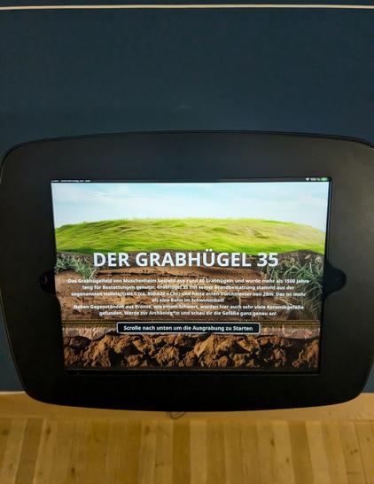 Ein festinstalliertes Tablet. Darauf sieht man den Schnitt durch einen Grabhügel. Darüber liegt in weißer Schrift "Grabhügel 35" und der Hinweis "Scrolle nach unten, um die Ausgrabung zu starten".

Außerdem wird in Schriftform erklärt, dass das Gräberfeld Muschenheim mehr als 1500 Jahre lang genutzt wurde und aus 40 Grabhügeln besteht. Nr. 35 stammt aus der sogenannten Hallstattzeit C, also circa 800-620 vor Christus.