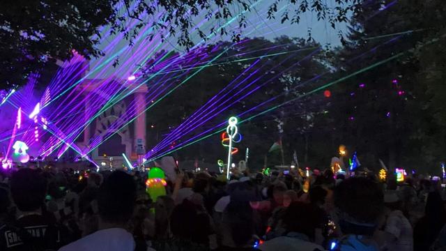 A crowd of people gathered around a brightly lit stage with purple and green laser lights shining above. Many people in the crowd have various glowing objects.