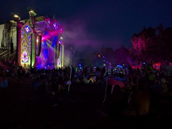 A large and brightly lit stage can be seen on the left, while fog coming from the stage is illuminated by purple, yellow and blue lights.  There is a crowd of festival goers around the stage.