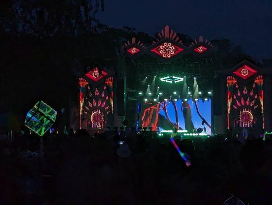 A brightly list show stage with vibrant red, yellow and green patterns.  A large crowd is gathered in front of the stage with various glowing objects.