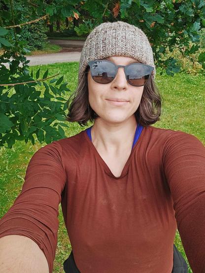 A colour photograph of a woman who is wearing a brown wool hat and black sunglasses and smiling very slightly. Around her is trees and grass.