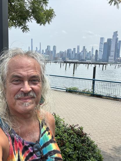 A selfie of a white haired man, with the Hudson river & midtown Manhattan behind him.