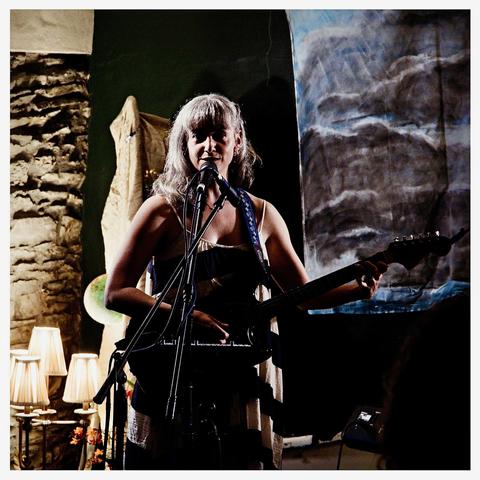 Inni-K at the mic looking towards us with her guitar. A backdrop by Bee hangs of to her left, a brooding cloud and ocean scene. Three little lamps illuminate a stone wall to her right.