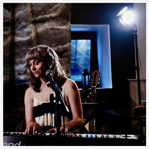 Left of frame, Inni-K at the mic eyes closed playing her Roland keyboard. Over her left shoulder we see a standing guitar and a window. Inthe topright a small light on a stand. More of the backdrop by Bee hangs to her right, a brooding cloud and ocean scene.