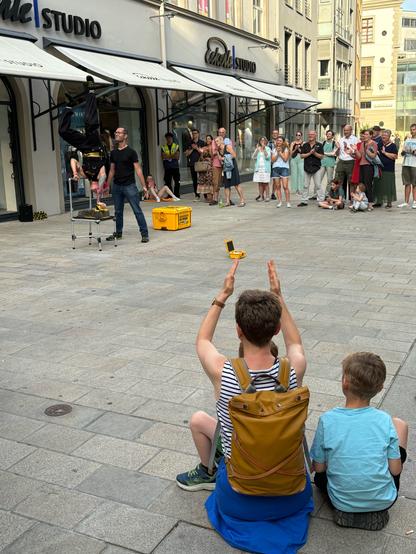 In einer Einkaufsstraße in Augsburg mit modernen Geschäften findet eine Straßenperformance statt. Eine Akrobatin hängt kopfüber auf einer Vorrichtung mit einem Nagelbett unter ihrem Kopf, während ein Assistent daneben steht. Die Zuschauer, darunter Erwachsene und Kinder, klatschen und beobachten die Darbietung aufmerksam. Ein Kind und eine Erwachsene sitzen im Vordergrund auf dem Boden und applaudieren.