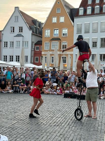 Auf dem gepflasterten Rathausplatz in Augsburg, umgeben von farbenfrohen historischen Gebäuden, findet eine Vorführung statt. Ein Mann auf einem hohen Einrad wird von einem Assistenten gestützt, während eine Frau in roter Kleidung neben ihm steht. Das Publikum, bestehend aus Erwachsenen und Kindern, sitzt und steht rund um die Darbietung und schaut gespannt zu.