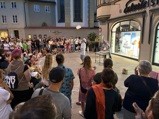 Eine große Gruppe von Menschen steht und sitzt in einer belebten Fußgängerzone vor einem Schaufenster. In der Mitte der Szenerie stehen zwei Personen in Kochkleidung. Einer der Köche wirft Teig in die Luft, während die Menge zuschaut. Das Publikum besteht aus Erwachsenen und Kindern, die aufmerksam dem Geschehen der beiden Clowns folgen und applaudieren.