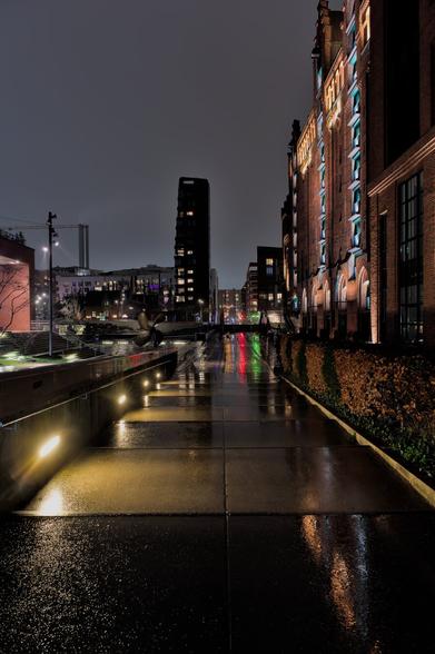 A square with a long, flat staircase in the urban environment. The ground is wet. Surrounding the square are buildings with lighting in various colors. The stairs are also illuminated. On the right is an old, illuminated warehouse building.
The whole scene is reflected in the wet ground.