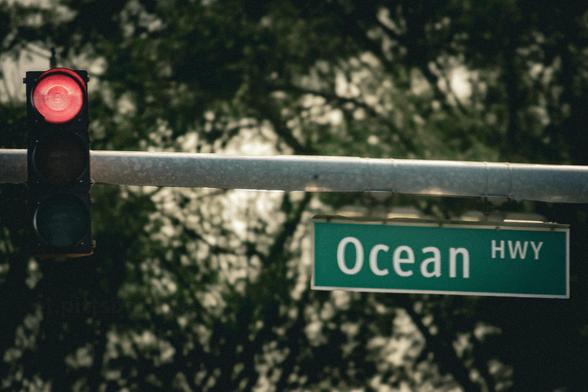 Traffic light showing red next to a green road sign reading "Ocean HWY" with trees in the background.