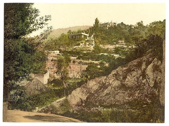 The image depicts a picturesque landscape scene with a mix of natural and human elements. In the foreground, there is a stony path that winds through a lush green hillside dotted with trees and bushes. To the right, part of a house or a small villa is visible, set amidst the verdant surroundings. The house features a tiled roof and possibly has some architectural details visible, but they are not clearly distinguishable due to the distance from which the photo was taken.

The colors in the image are muted with soft earthy tones dominating the scene, giving it an old-fashioned or vintage feel. The overall composition and style suggest that this is a historical photograph from around 1900, showcasing the beauty of European landscapes during that time period.

The text provided includes additional information about the location—Menton, France—and the style of the image (photochrom prints), which are both relevant for understanding the context and historical significance of this picture.