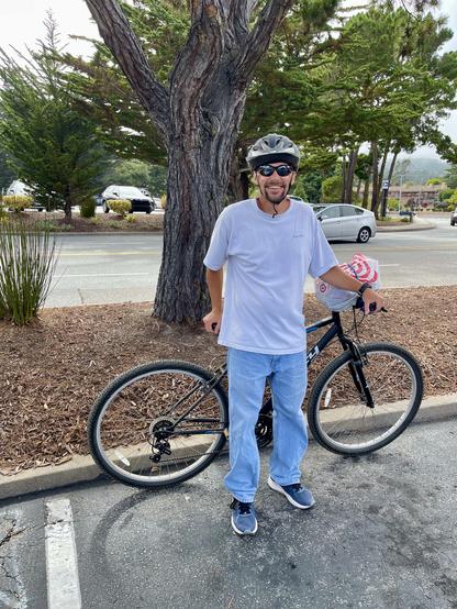 A smiling man stands in front of his bicycle in a parking lot near an entrance to Del Monte Center in the city of Monterey. He is wearing sunglasses, a gray/silver bike helmet, light blue denim pants, a gray t-shirt, and blue tennis shoes. Behind him are trees and some cars.