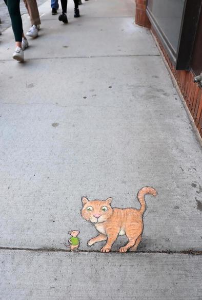 Streetart. A small cat and a mouse (named Eugene) were painted with chalk on a concrete sidewalk next to stores. The expansion joints were integrated into the picture. The orange and beige cat balances on the line next to the mouse in the green T-shirt. The mouse rests his arms on his hips and looks at the cat. Title: "Eugene's morning commute is disrupted once again by an unexpected game of mouse and cat."