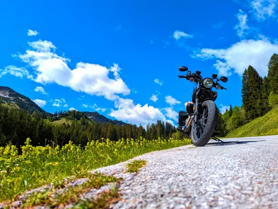 Das Bild zeigt eine malerische Berglandschaft unter einem strahlend blauen Himmel mit einigen weißen Wolken. Im Vordergrund steht ein schwarzes Motorrad auf einer asphaltierten Straße, die leicht ansteigt. Das Motorrad ist prominent im Bild platziert, während die Kamera einen tiefen Winkel verwendet, um es zu fotografieren. Die Reifen des Motorrads sind deutlich sichtbar, und das Design wirkt modern und robust.

Im Hintergrund erstreckt sich eine üppig grüne Landschaft mit dichten Nadelwäldern, die die Berghänge bedecken. Weiter hinten sind die Gipfel der Berge zu erkennen. Die Szenerie vermittelt ein Gefühl von Freiheit und Abenteuer, das mit einer Motorradtour in solch einer beeindruckenden Naturkulisse verbunden ist.

Im unteren Bereich des Bildes sind auch einige Wildblumen und Gräser zu sehen, die die Straße säumen und dem Bild zusätzliche Farben und Texturen verleihen. Die klare Luft und das helle Sonnenlicht tragen zur lebendigen und frischen Atmosphäre bei. Insgesamt fängt das Bild die Schönheit und die Ruhe der Natur ein, kombiniert mit dem Abenteuergeist des Motorradfahrens.