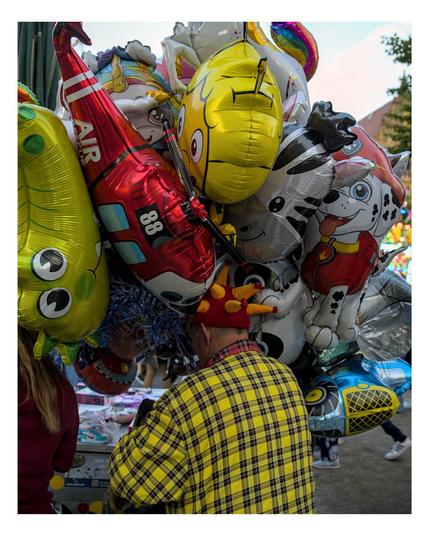 bunt verkleideter Luftballonverkäufer steht mit dem Rücken zum Fotografen und ist mit seinen Ballons beschäftigt.