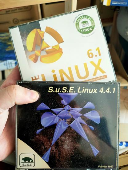 A hand holding two CD covers.
The one on the front is dark with a blueish structure on it. It's title says "S.u.S.E. Linux 4.4.1".
The one in the back is light with a yellow structure on it. Readable is only "Linux 6.1"
