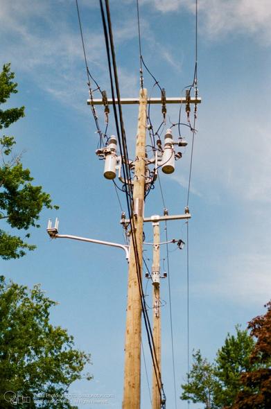 A photograph of two power poles with multiple transformers and wiring. On the frontmost pole, about halfway up, a diagonal metal pole emerges from the left side holding aloft a metal box with four short antennae on top. Beyond are some trees and a blue sky with some wispy clouds.