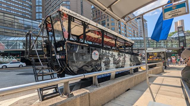 The image shows a Boston Duck Tour vehicle, which is an amphibious bus used for city tours in Boston. The vehicle is black with an open top and a canopy. It is parked at a designated "Duck 1 Boarding Area" next to a curb on a city street, with a sign indicating it is ready for boarding. The front of the bus features an American flag, and there are stairs for passengers to board. The website "www.BostonDuckTours.com" is prominently displayed on the top front of the bus. In the background, tall buildings and a pedestrian bridge are visible. A small kiosk selling tickets and souvenirs is seen to the right, with people walking nearby.