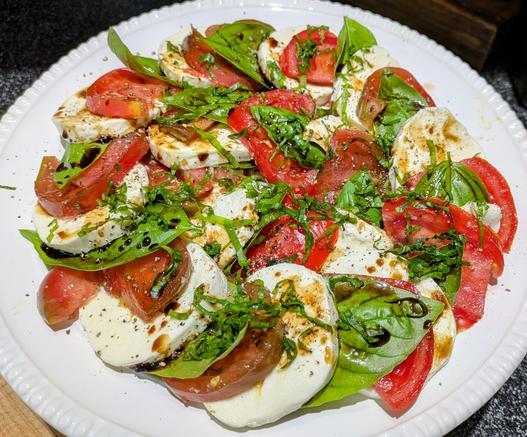 A large plate of caprese salad with mozzarella basil heirloom tomatoes and balsamic vinaigrette. A chiffonade of Basil is over top along with salt and pepper.
