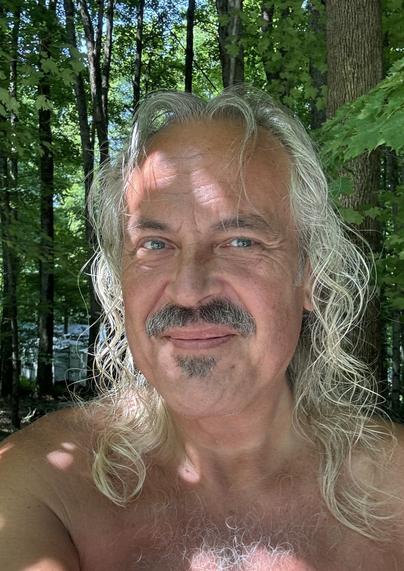 A white haired man in a selfie in the woods. Dappled light from the trees lights his head and shoulders, woods behind.