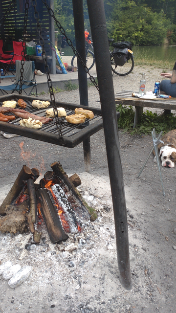 Ein Dreibein-Grill mit rechtwinkeligem, gut belegtem Grillrost. Rechts eine scheckige Bulldoge am Boden liegend. Im Hintergrund Velos und eine auf einer Seite offene Hütte.