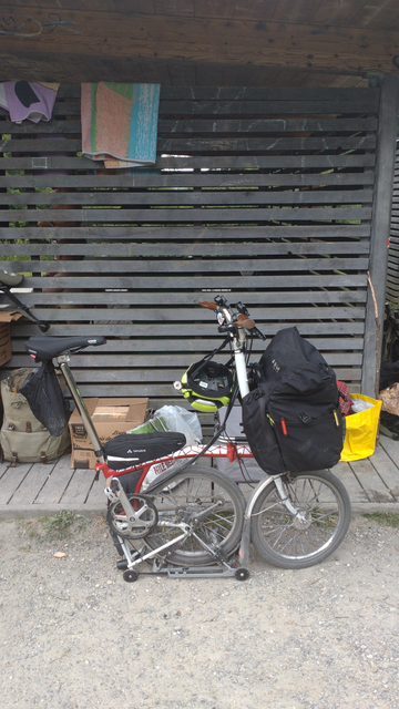 Ein halb gefaltetes, rot-weisses Brompton-Faltrad mit offener Fronttasche vor einer Holzwand mit etwas Gepäck und Brennholz an der Wand stehend.