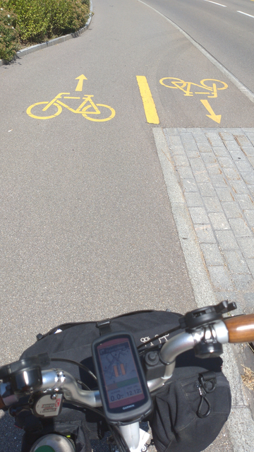 Gehweg auf der linken Seite einer Strasse. Fahrbahnmarkierungen zeigen, dass Velofahrer in Blickrichtung links fahren sollen und Velofahrer in Gegenrichtung auf der (in Blickrichtung) rechten Seite runter auf die Fahrbahn.

Im Vordergrund der Lenker und die Fronttasche eines Brompton-Faltrades.