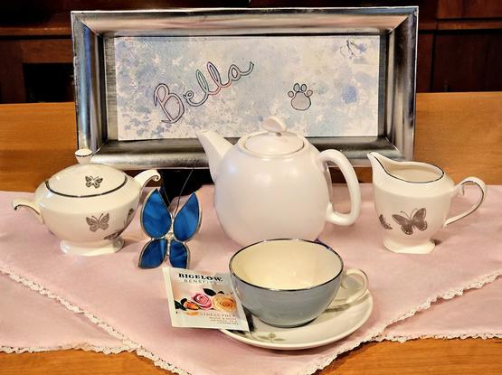 A tea tableau in pink & silver with hints of blue, and a theme of butterflies flying freely. There is also a sign showing the name "Bella" and a pawprint set against a sky. This is a memorial for a friend's dog. The tea is called Stress Free, a rose & mint herbal from Bigelow.