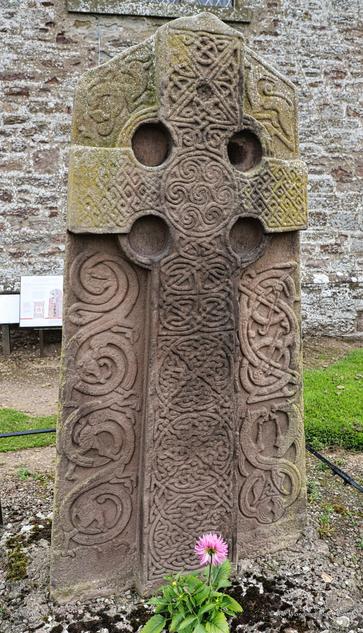 A photo of the graveyard stone cross, with beautiful and intricate Pictish carving of swirls, plaits and knots. The slab is rectangular but with a pointed top, and the cross itself is carved in bas relief. The areas surrounding the cross depict mythical creatures and windings. The stone itself is brownish-grey, with age-worn faces and lichen growing in spots.