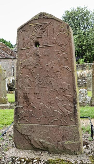 A photo of the obverse side of the stone depicting a battle. Armed knights on horseback and foot soldiers with swords, bows and spears are facing each other, and one figure - presumed to be the defeated king - is being attacked by a raven, usually indicating that he perished. The top of the stone is intricately carved with runes, the meaning of which isn't clear.