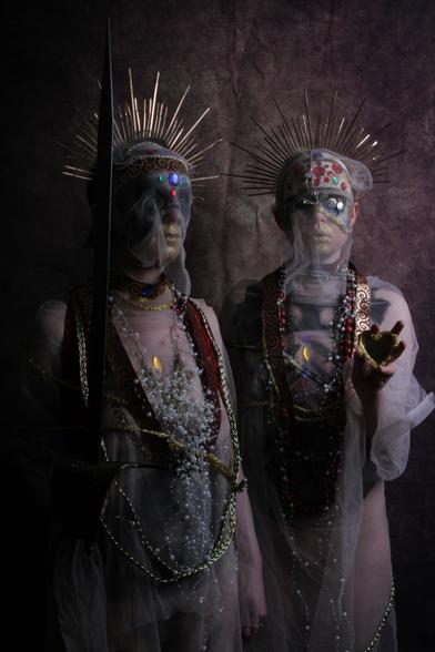 Two figures stand close together in semi-darkness lit by a single light source above them to the right. The figure on the left is at a 45 degree angle to us and holds a sword upright in their right hand, the figure on the right is facing us directly and holds a small heart-shaped mirror in their left hand, directly pointed at us.

Both figures are draped in gauze-like material, wearing headdresses in the forms of sun crown halos, with golden rays forming semi-circles above their heads. Beneath the gauze the top half of their faces/heads are covered with jewel-encrusted masks, and their lower faces seem golden. They have jewels, necklaces, and embroidered ribbons of cloth draped around their necks and covering their chests.