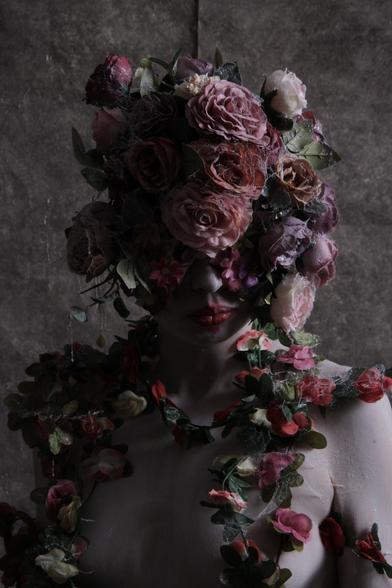 A figure’s head and shoulders are shown against an indistinct background. The figure’s head and top half of their face is covered with a mask/headdress made of faded pink, purple, white, and lavender roses. Some of the petals are covered with traces of cobwebs.

The figure’s lips are painted red, and their skin is alabaster. They have further strings of flowers around their neck, hanging down over their chest, similarly covered in cobwebs.