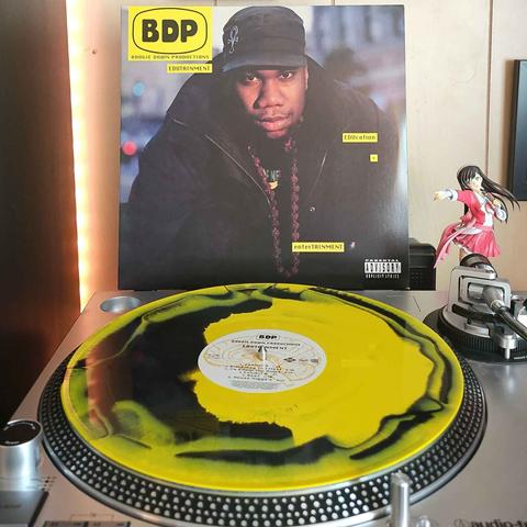 A vinyl Black Opaque & Canary Yellow Splatter record sits on a turntable. Behind the turntable, a vinyl album outer sleeve is displayed. The front cover shows KRS One wearing a heavy jacket, hate and beads while looking at the camera.