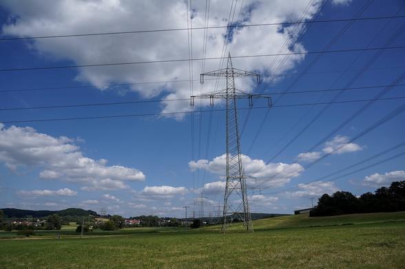 Das Bild zeigt eine ländliche Landschaft mit grünen Wiesen und einem blauen Himmel, der von weißen Wolken durchzogen ist. Im Vordergrund und entlang der Landschaft sind mehrere Hochspannungsleitungen und Strommasten sichtbar, die sich durch die gesamte Szene erstrecken. Die Strommasten sind aus Metall und haben eine typische Gitterstruktur. Im Hintergrund sind Bäume, Hügel und ein kleines Dorf mit Häusern zu erkennen. Die Umgebung wirkt ruhig und idyllisch, wobei die Präsenz der Stromleitungen die Verbindung zur modernen Infrastruktur verdeutlicht.