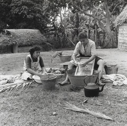 This image depicts a scene of traditional laundry practices. Two girls are seated on the ground, engaged in washing clothes. The girl on the left is using a large metal bowl filled with water and some detergent to scrub her clothes, while the girl on the right is using a washboard to clean hers in a larger metal tub. Both of them appear focused on their task. In the foreground, there's a large kettle, likely used for boiling water or steaming clothing. Behind them, a dog is drinking from another large metal tub, adding a casual and domestic atmosphere to the scene. The image captures a moment of everyday life in a location that seems to be a rural setting, possibly in a village or a community where traditional washing methods are still commonly used.