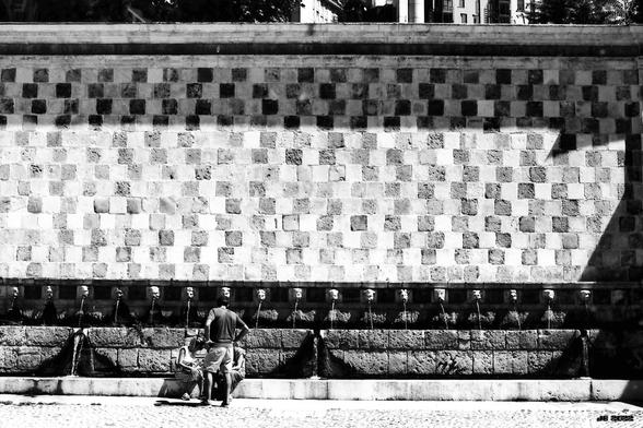 Schwarzweißfoto des Brunnens "Fontana delle 99 cannelle" in l'Aquila, Abruzzo, Italien. Dem Namen entsprechend umfasst die Brunnenanlage 99 Wasserspeier, von denen auf diesem Bild 18 zu sehen sind. Das Wasser tritt aus steinernen Köpfen aus. Über dem Brunnen eine Mauer aus Stein mit feinem quadratischem Muster. Leica R5 mit 50 mm Summilux auf Adox CMS II-Schwarzweißfilm.