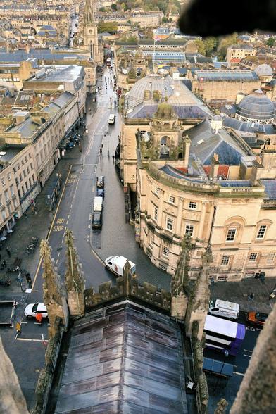 Looking down at street bordered by yellow stone buildings with black roofs.  In a top corner is a black shadow and at the bottom, either corner are edge of stone, from the parapet on top of the tower.

The most obvious building in the street is on the corner and have a curved facade. It's three storeys  high, judging from the three levels of windows (all multi-paned with white frames and dark glass, the upper level are smaller) Between the windows are bricks are ground level, and columns on the upper levels. One side is shaped like a tower, topped with a stone gazebo-like thing over a skylight. There is a similar one of the far side of the adjoining building, which has a central dome.
For all that the top of the roofs of buildings can be seen, there's not much sense of height.

In the foreground is large reverse-V shaped black roof with stone edging and two spires at the visible corners. The roof of the abbey building below.