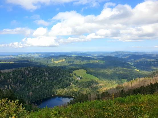Blick auf die Hügel des Schwarzwaldes, blauer Himmel mit Quellwolken, vorne tiefer unten ein kleiner See, in dem sich der Himmel spiegelt.