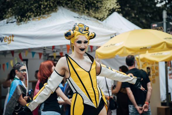 Drag Queen Vava Vilde im gelben Outfit und im Hintergrund der Stand von bwegt im Rahmen des CSD in Stuttgart.