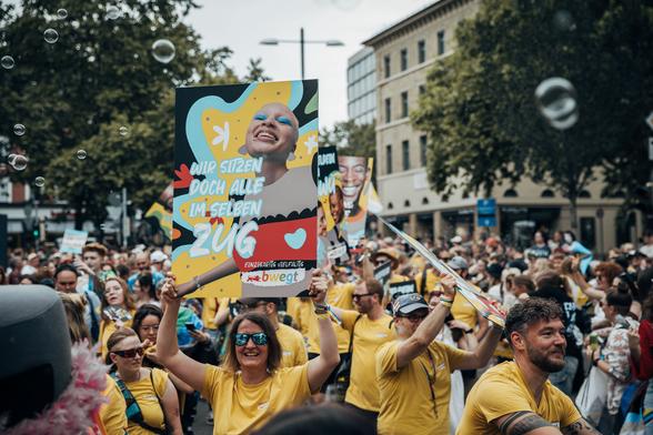 Menschen in gelben T-Shirts bei der CSD-Demo in Stuttgart, auf einem bwegt-Plakat steht: "Wir sitzen doch alle im selben Zug"